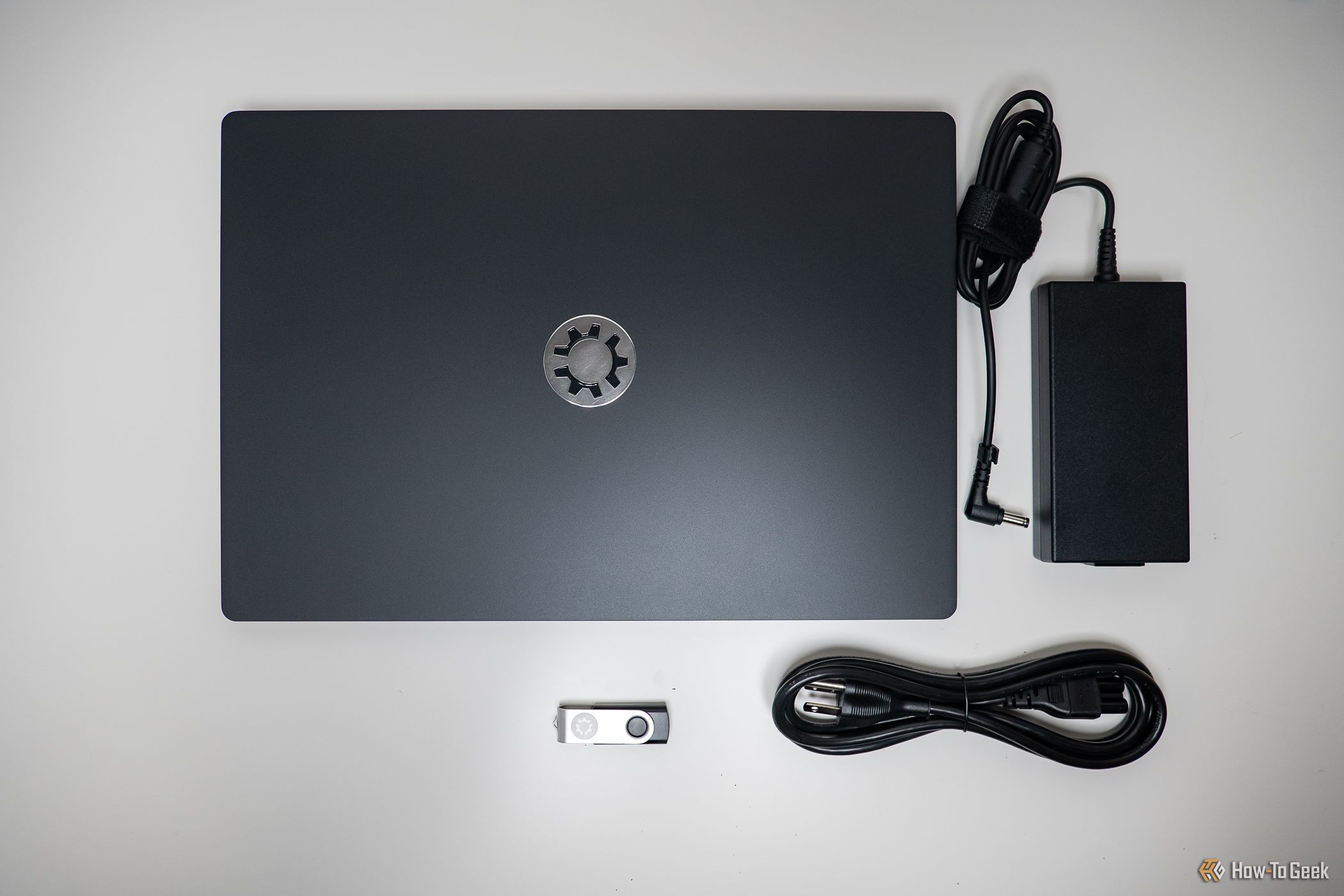 Top-down view of a closed Kubuntu Focus Ir16 Gen 2 laptop along with its PSU, wall plug, and factory reset USB drive.