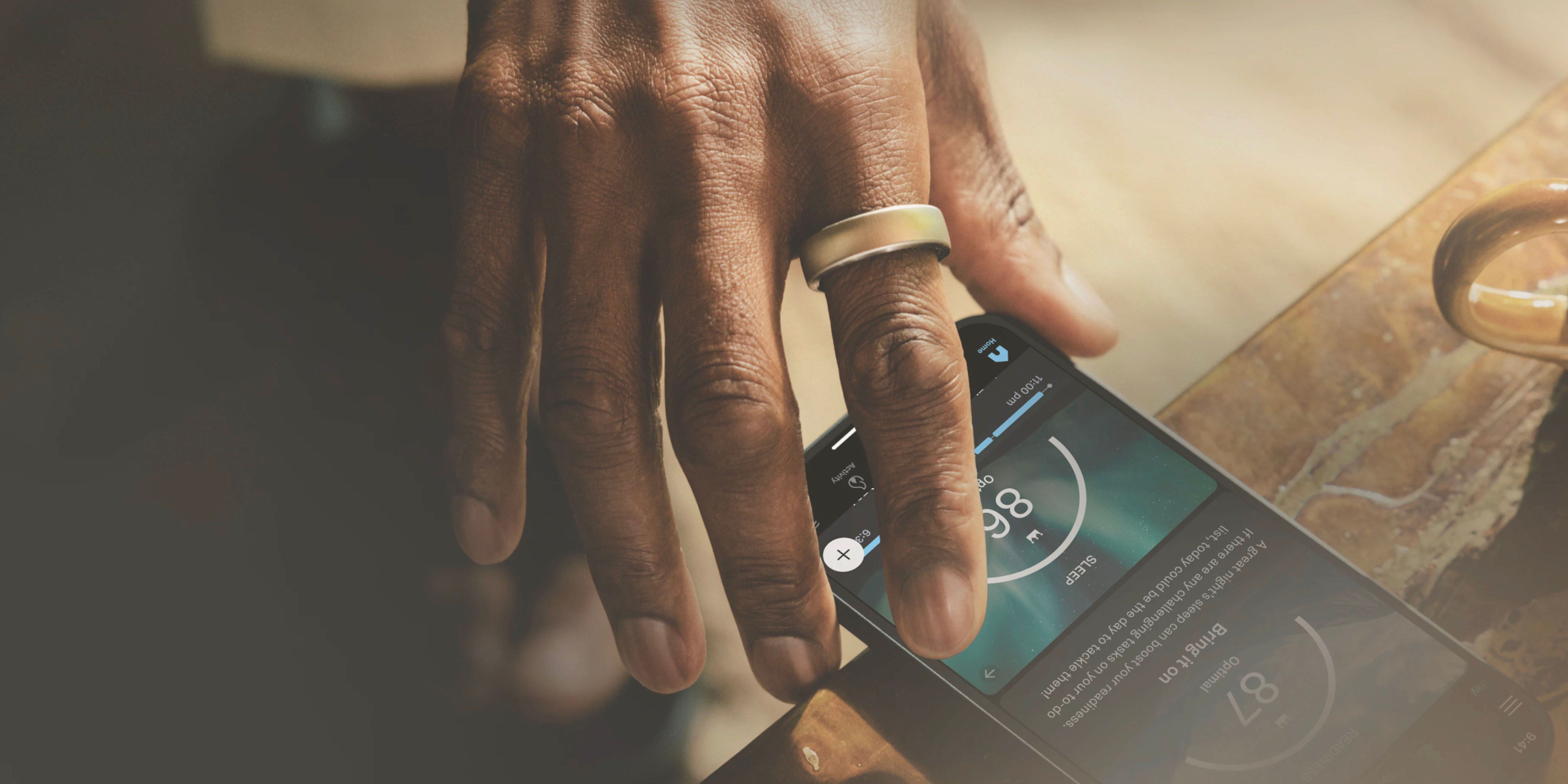 Oura promo shot of a hand wearing a ring, reaching for a phone with the app