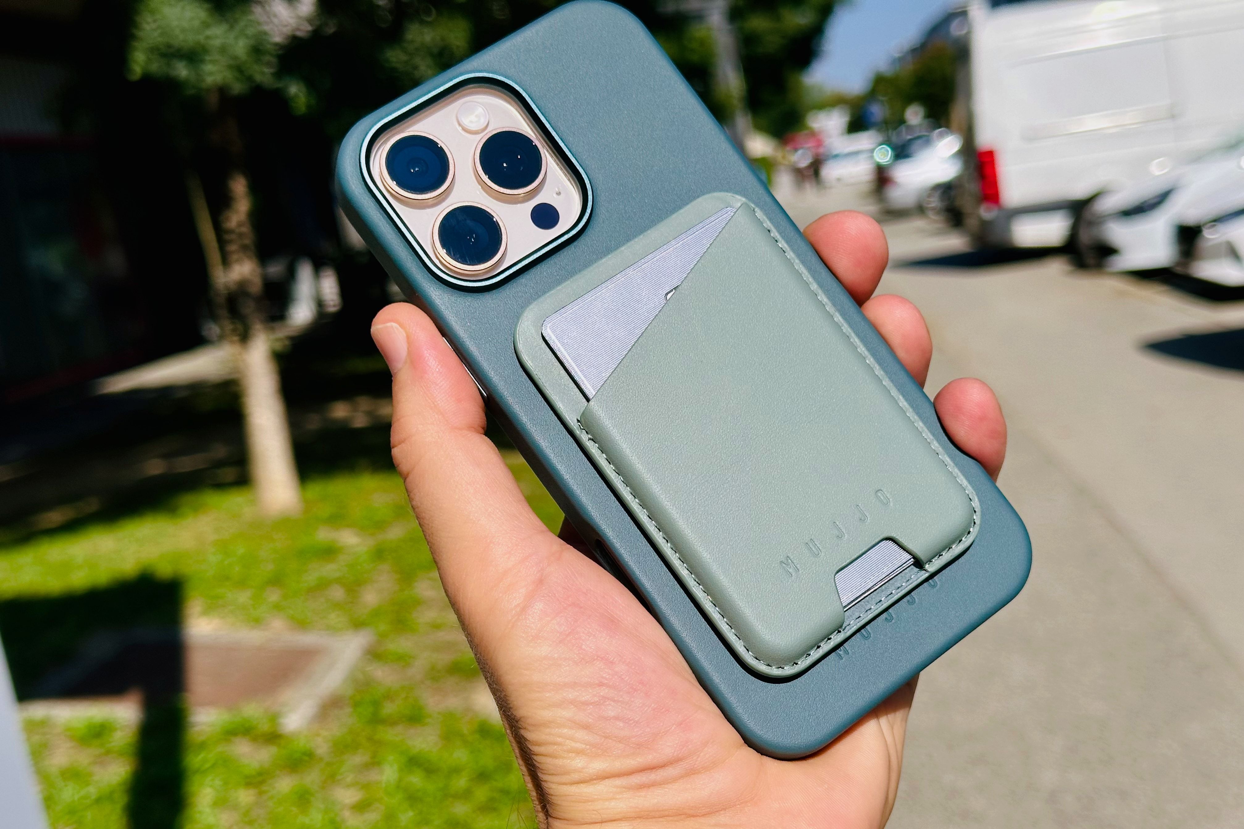 Hand holding an iPhone 16 Pro Max in a Mujjo leather case and MagSafe wallet, set against a blurred street in the background.