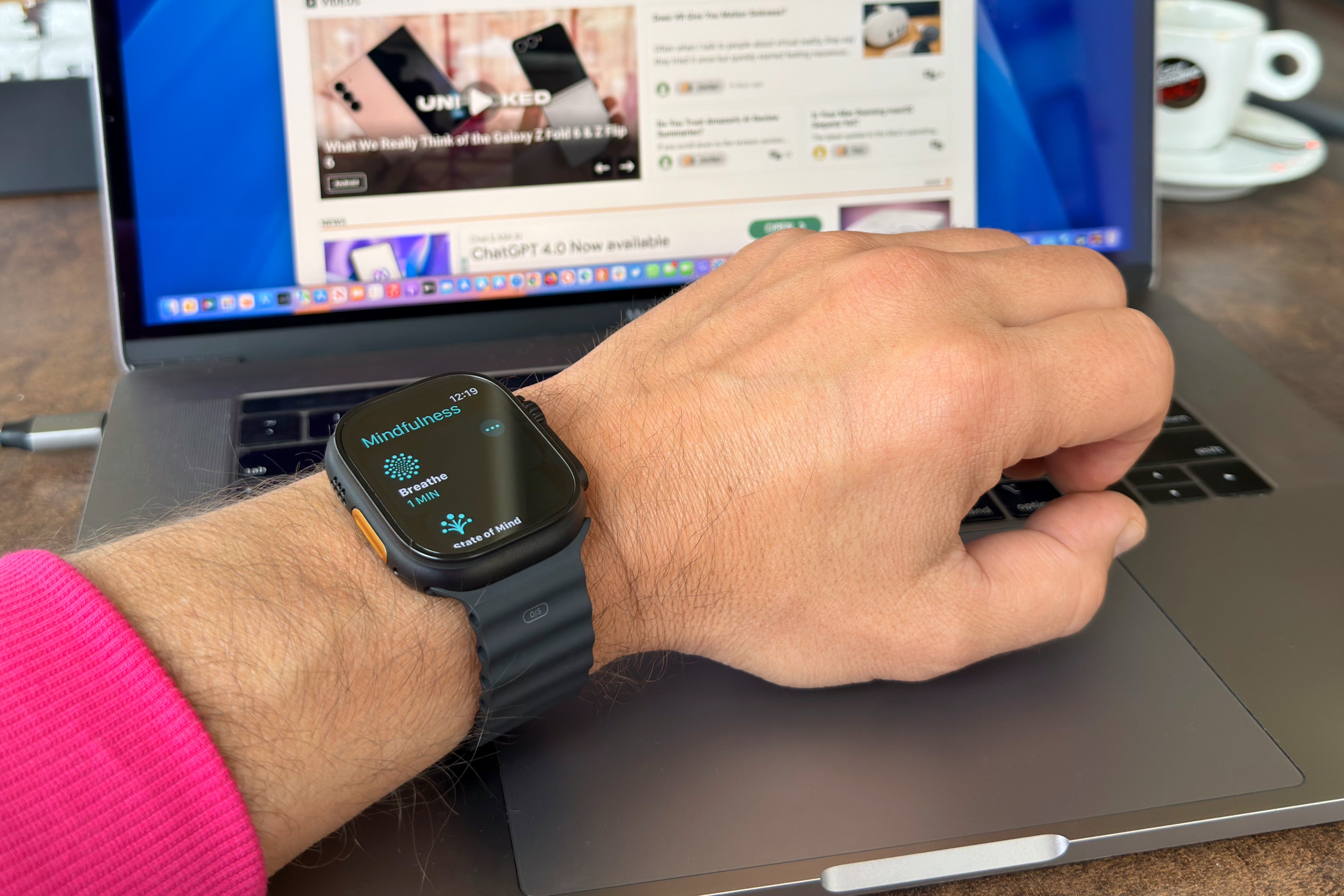 Person's hand resting on a MacBook Pro laptop. Apple Watch Titanium is on their wrist, displaying the Breathe option in the Mindfulness app.