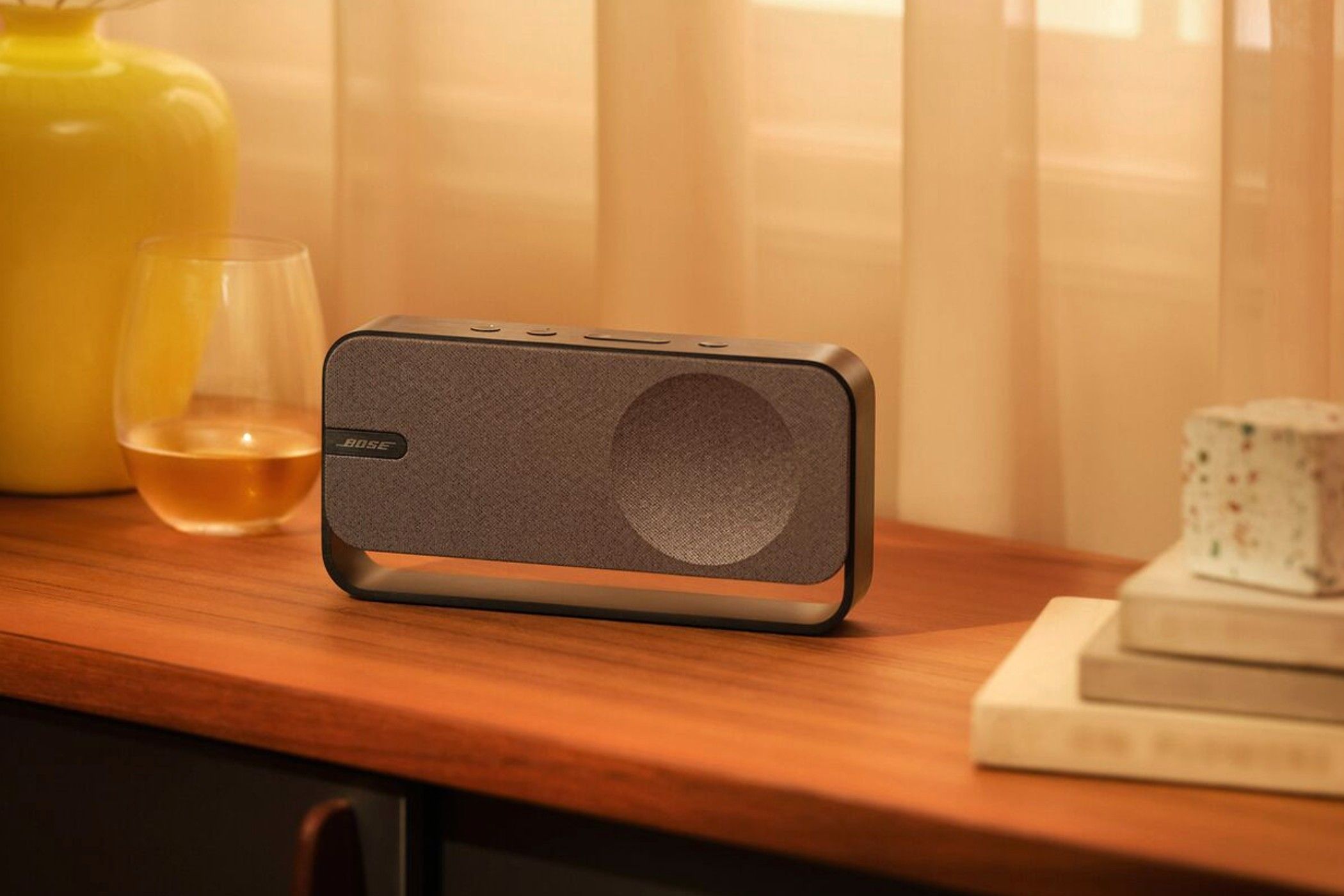 Bose SoundLink Home Bluetooth Speaker on a table between books and a glass of liquid