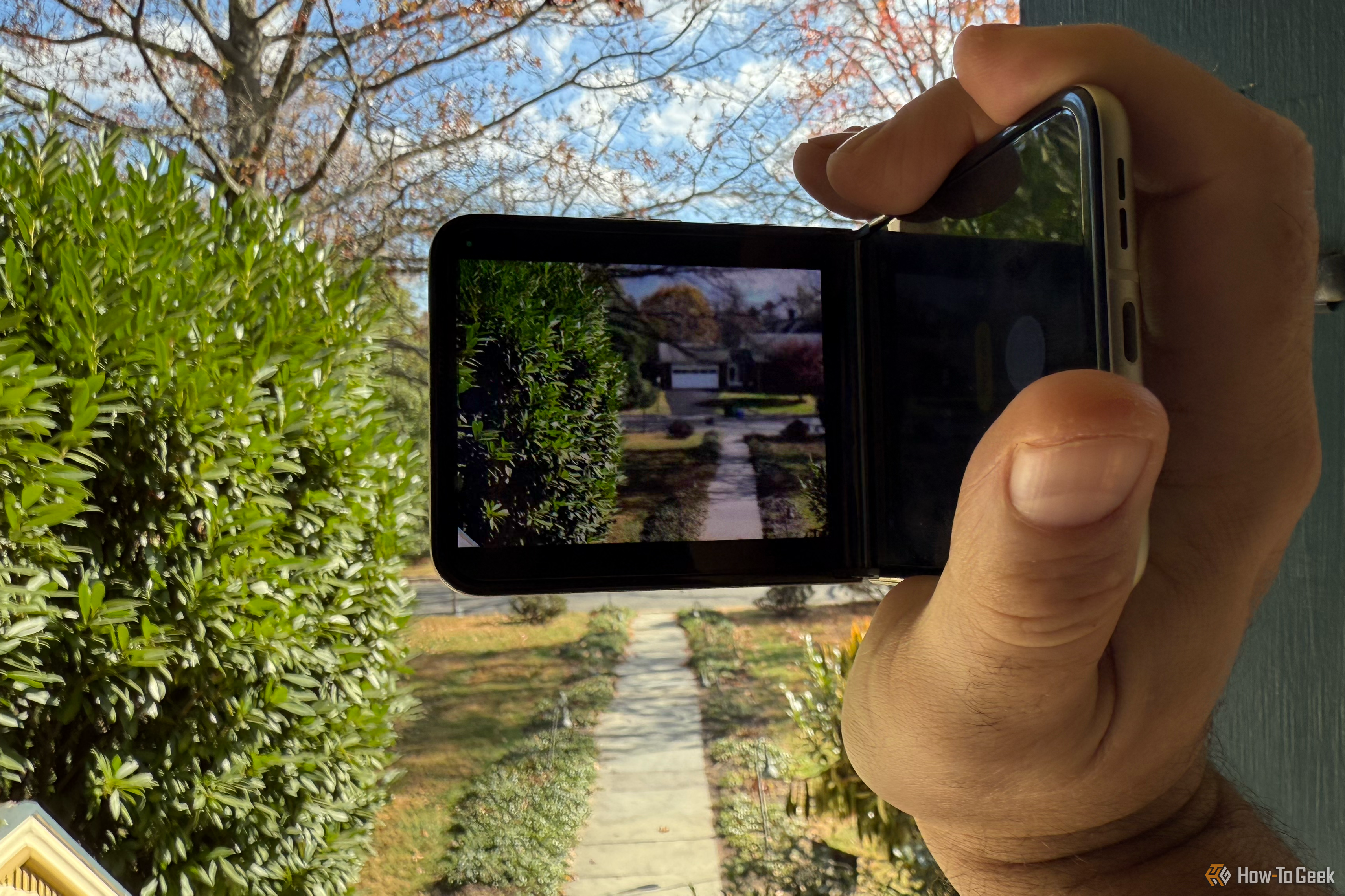 A hand holding the Motorola Razr at an awkward angle. Bushes and bright skies are seen on the display, and in real life right behind it.