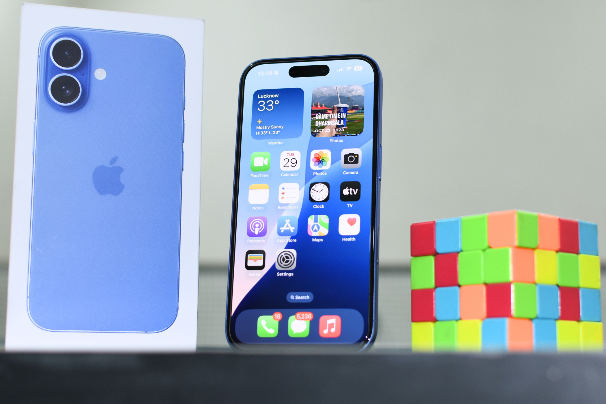 iPhone 16 on a shelf with its box on the left and a rubik's cube on the right.