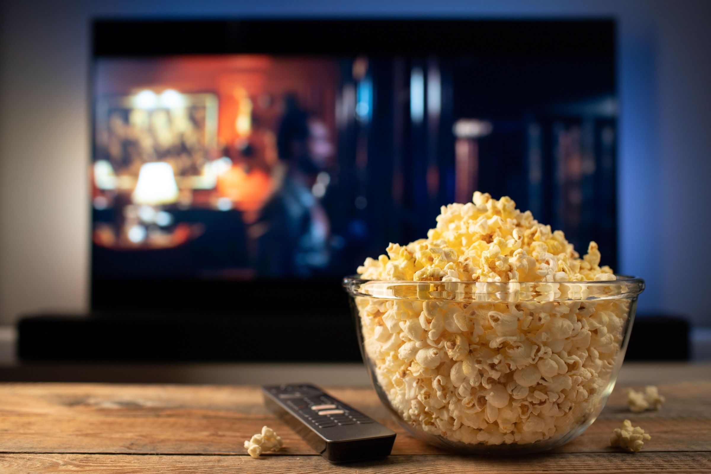 Shutterstock_2256308249A glass bowl of popcorn and remote control while in the background the TV plays.