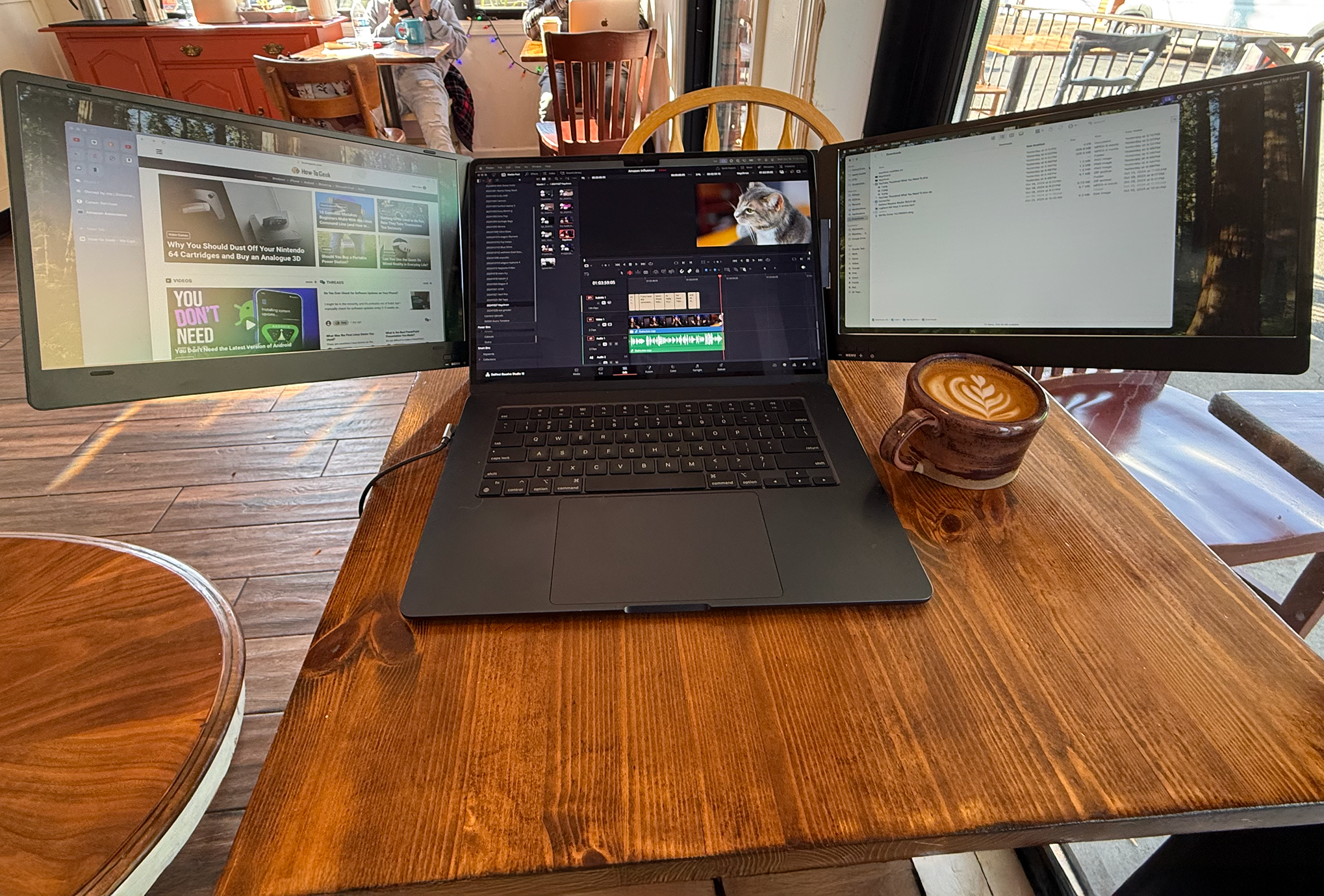 A MacBook Air sits on a table in a coffee shop with the laptop flanked by two external displays. A coffee cup sits on the wood grain table.