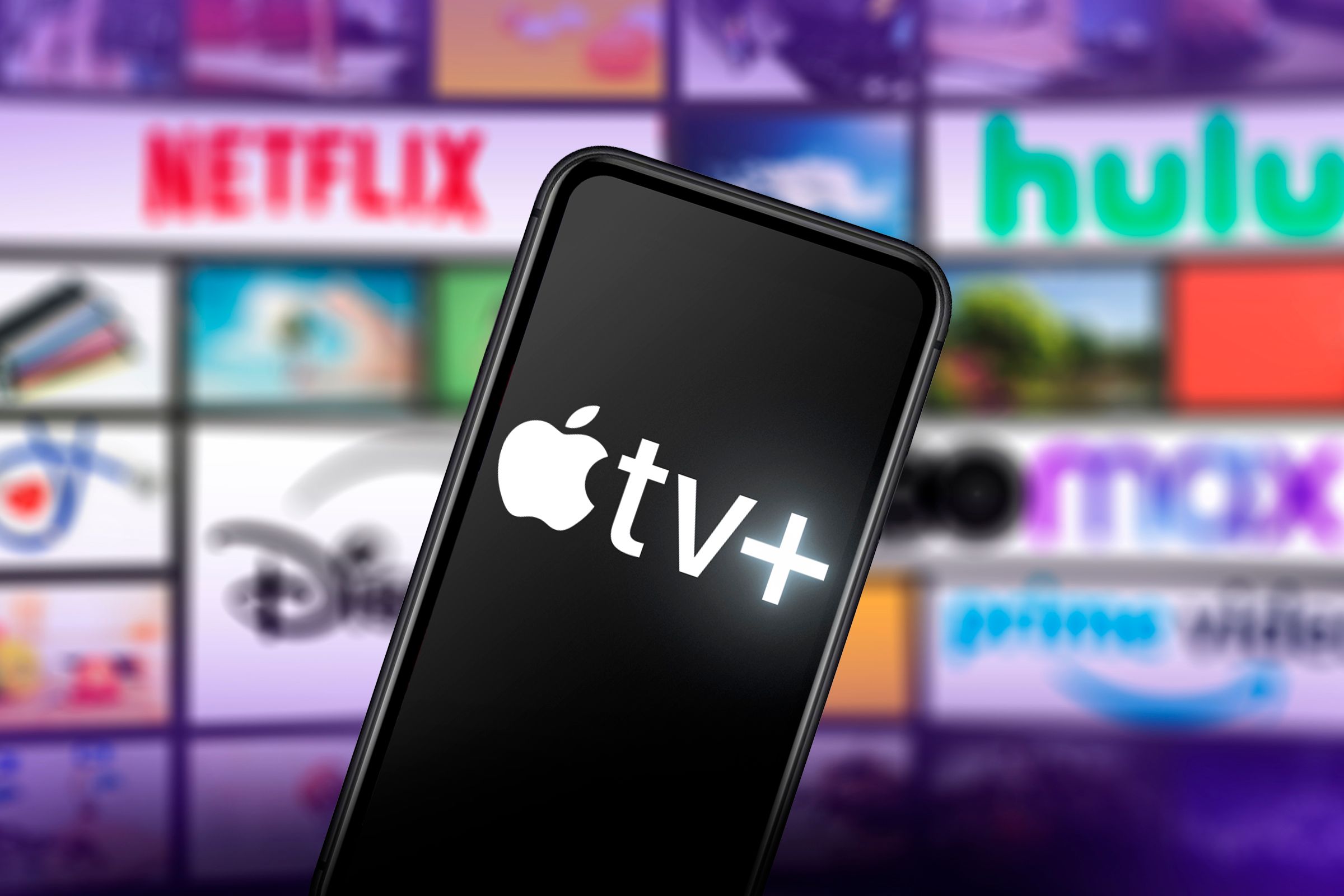 A phone with the Apple TV+ logo on the screen and in the background several screens with logos of other streaming services.