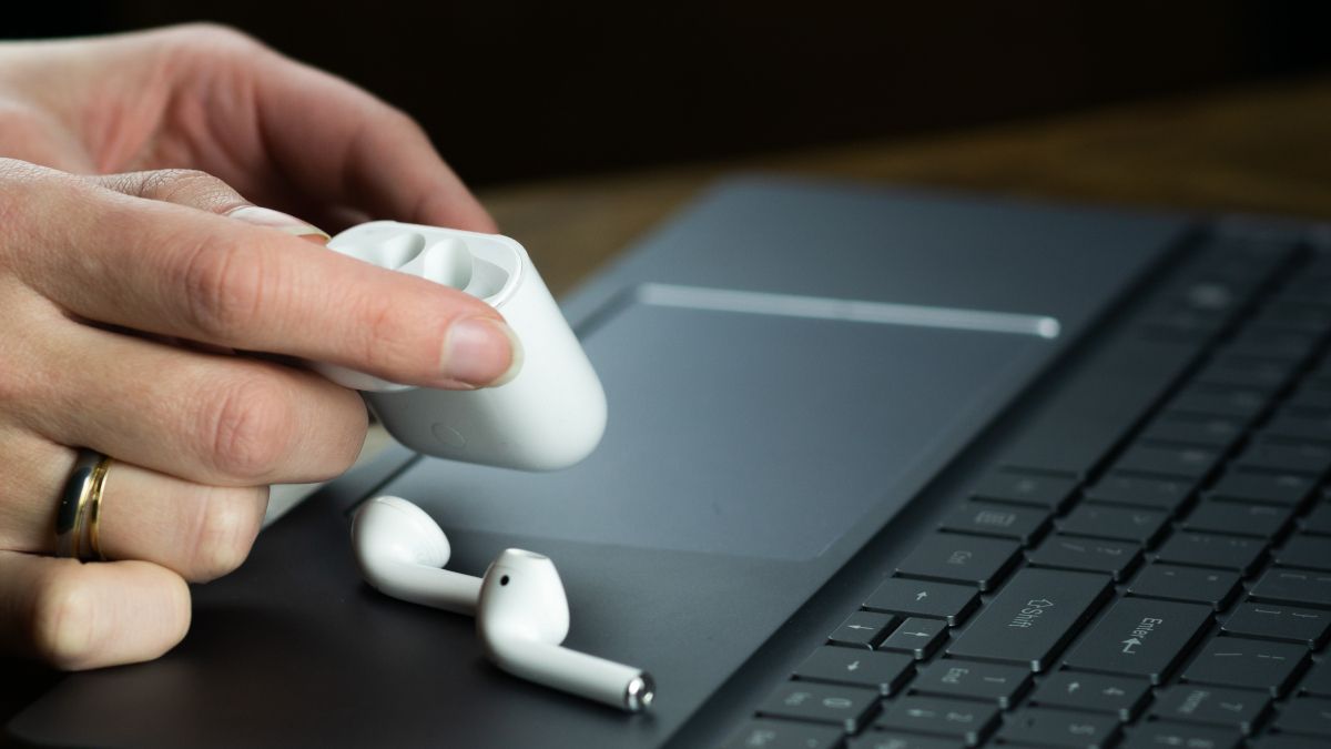 Person's hands holding an Apple AirPods case with the earbuds on top of black laptop.