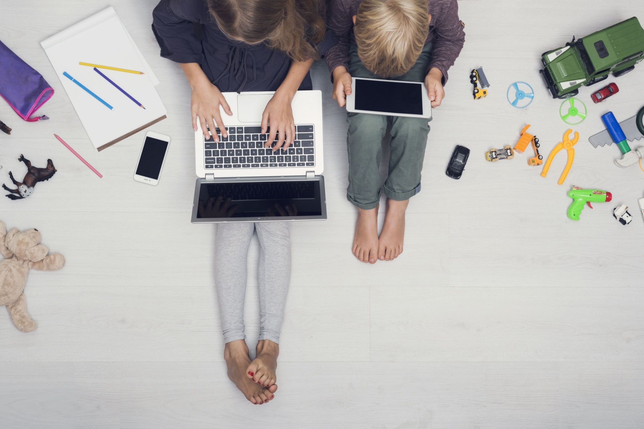 A girld with a laptop and boy with a tablet seen from above, surrounded by toys.