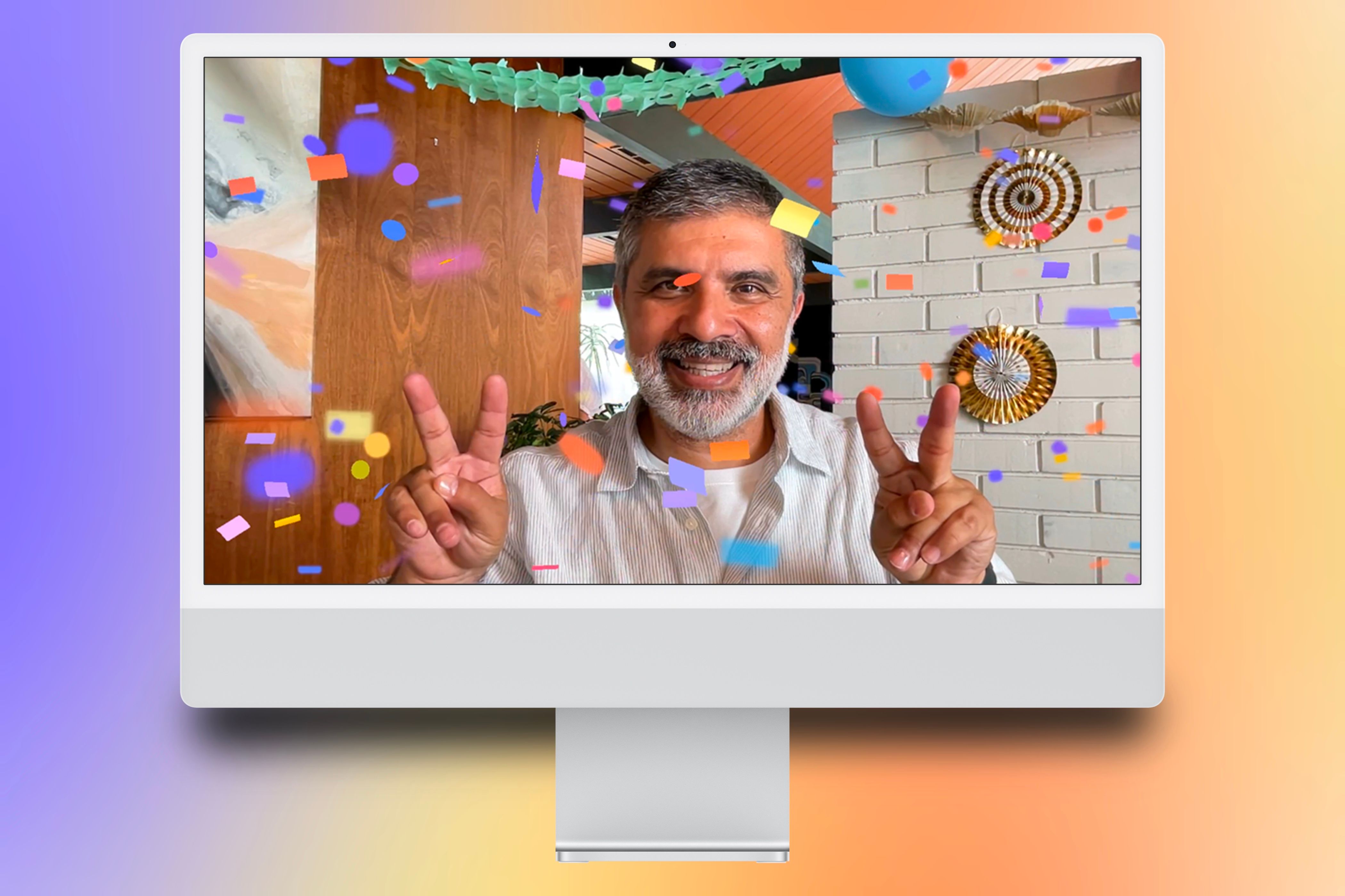 An iMac showcasing a person doing two peace-signs to enact the confetti reaction on a FaceTime call centered with a colorful background behind it.