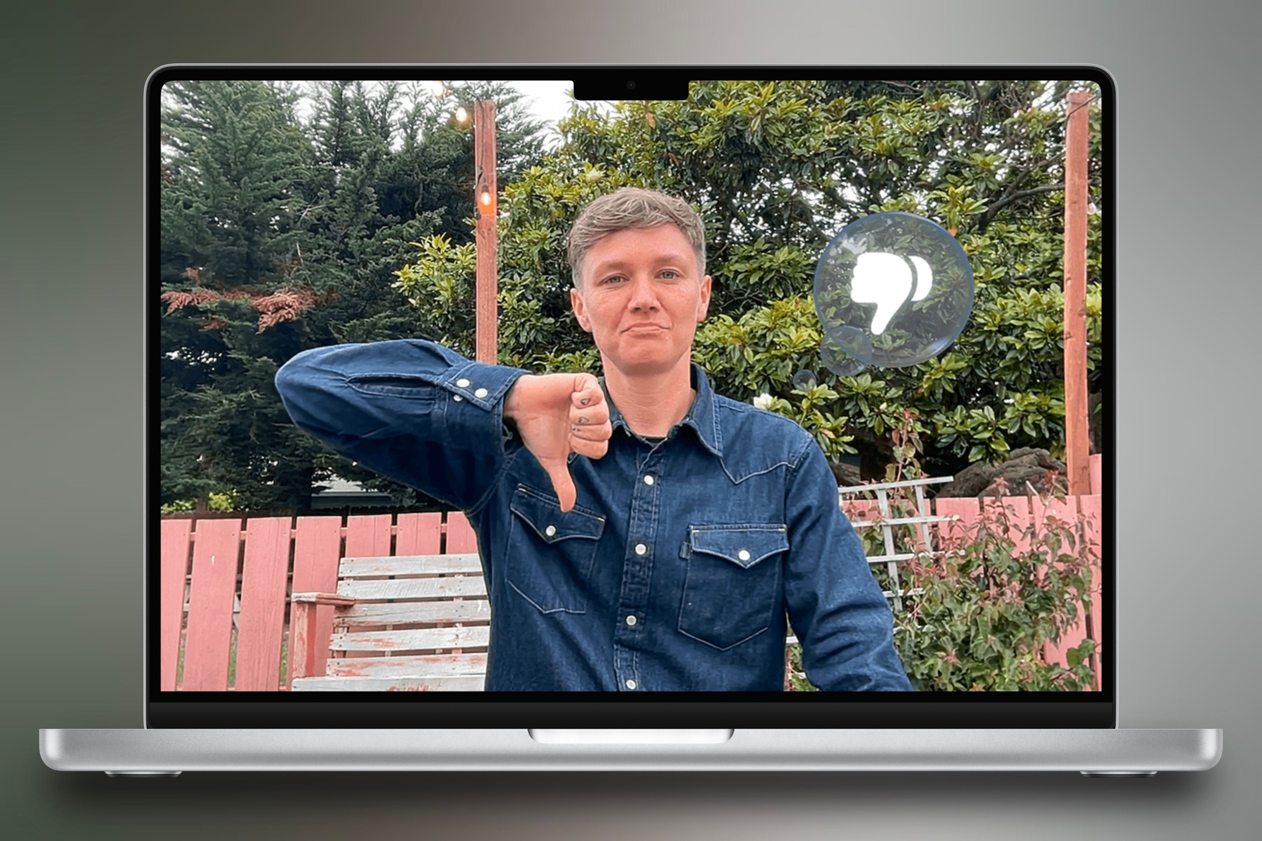 A MacBook Pro centered with a colorful background behind it showcasing a person giving a thumbs down while the corresponding reaction showcases on the call.