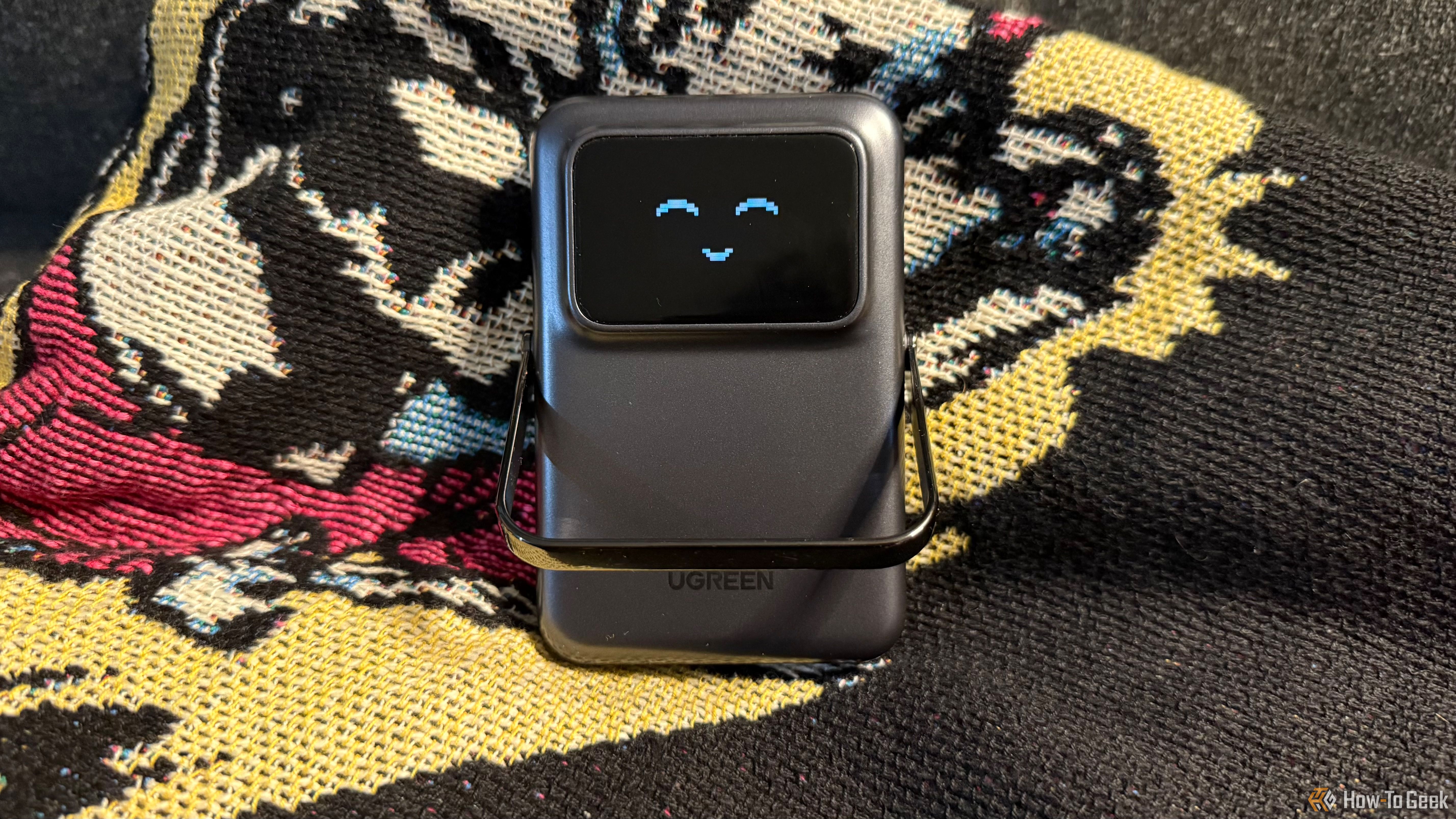 The Ugreen Uno Wireless Power Bank sitting on a blanket with a smiling emote on its screen and its adjustable stand down.