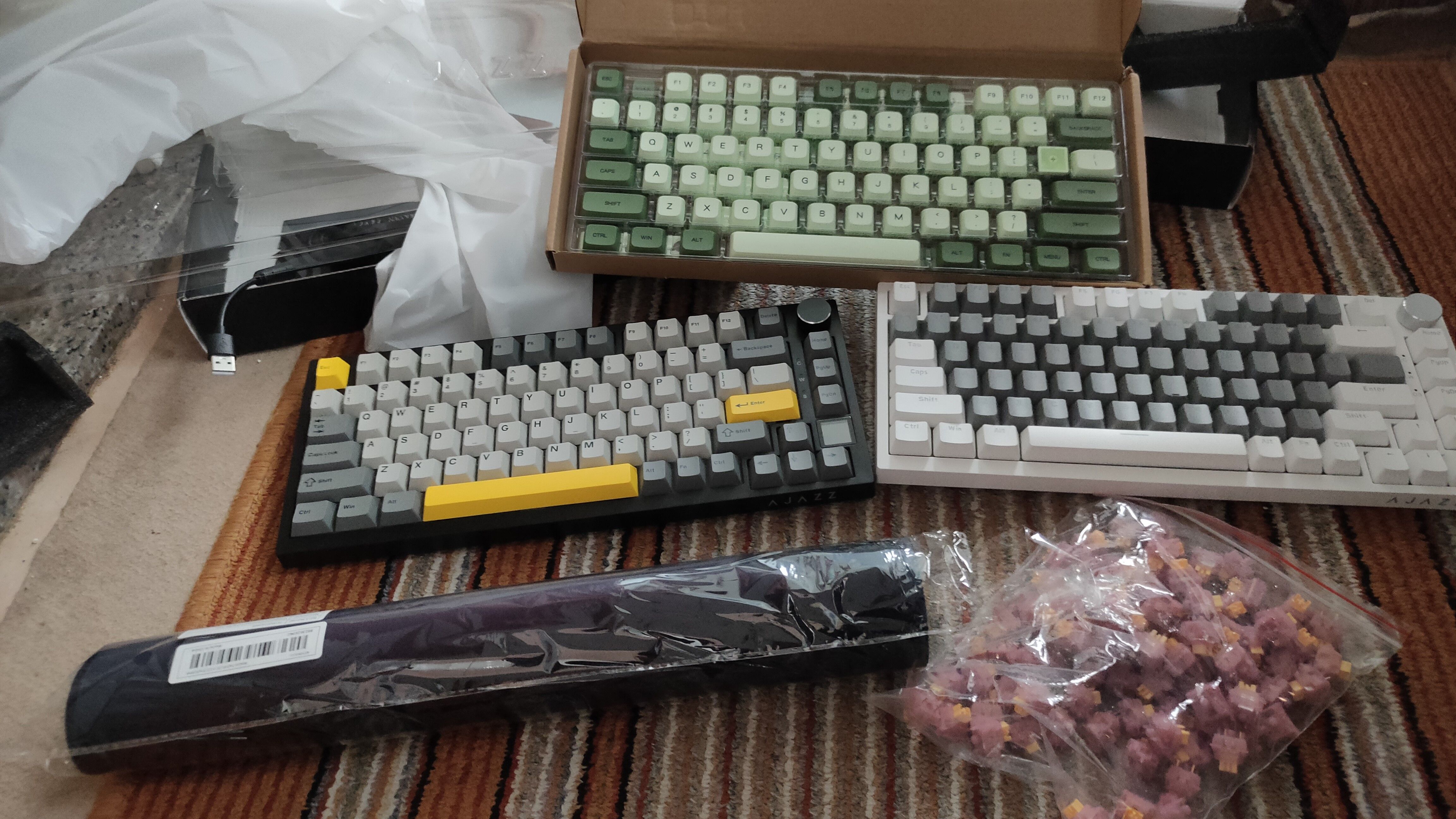 The Ajazz AK820 Pro and non-pro keyboards, along with a set of green keycaps, a rolled up mouse pad, and a bag full of Outemu Peach V3 switches on the floor.