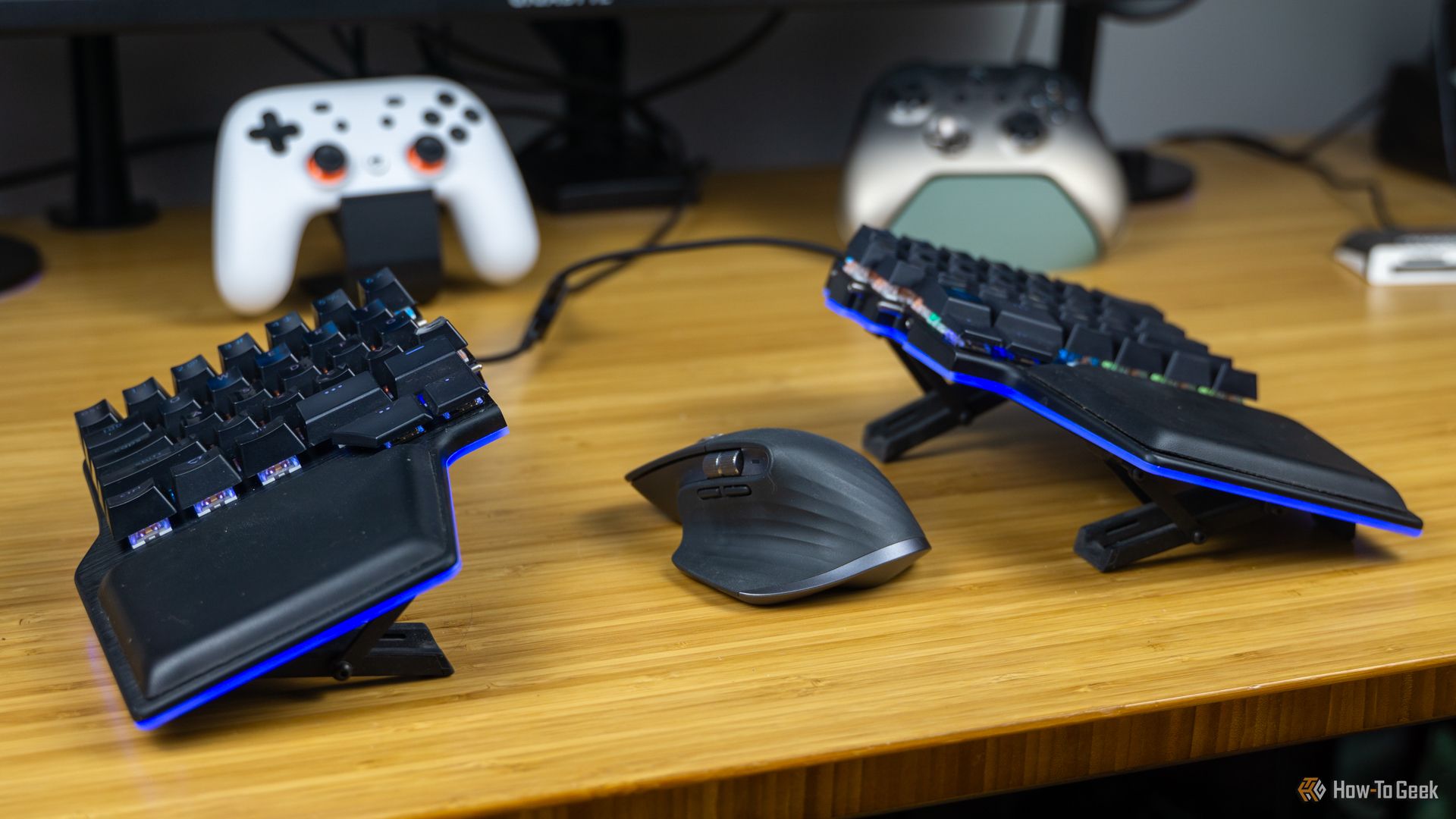 The Dygma Raise split keyboard on a bamboo desk with a MX Master 3 between the halves and some game controllers in the background.