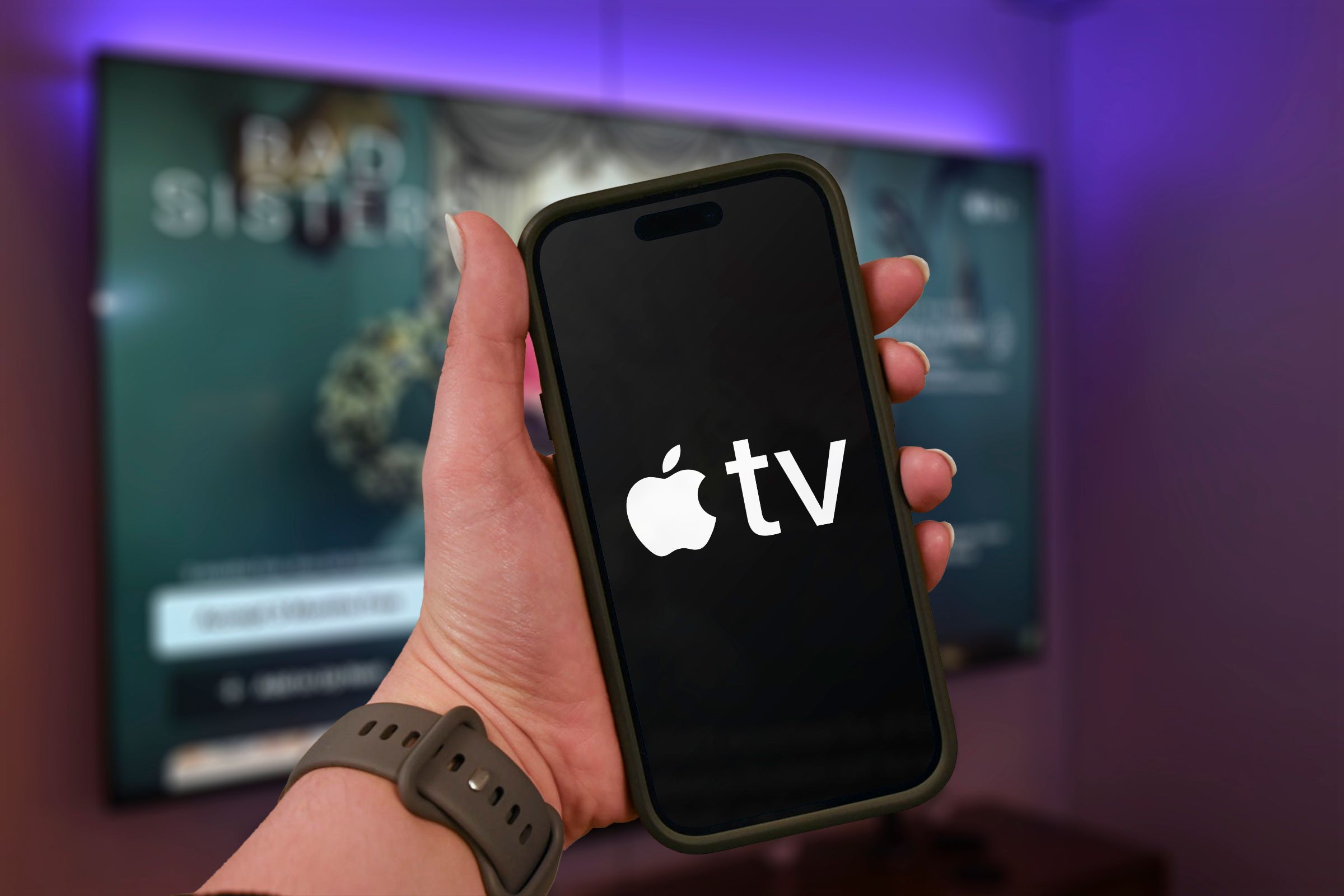 Hand holding a phone with the Apple TV+ logo and a blurred TV in the background.