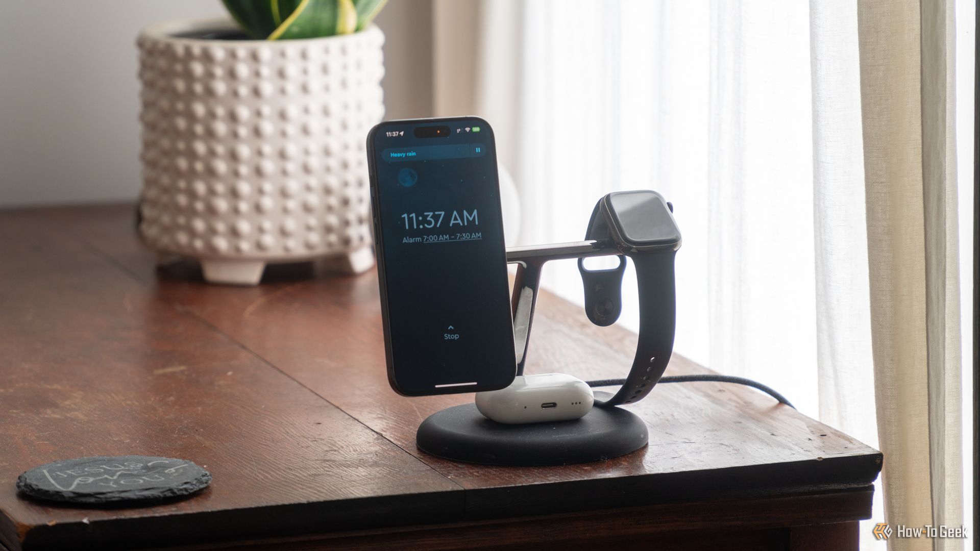 The Sleep Cycle app with an alarm set showing the wake up window on an iPhone 15 Pro docked on a charger with an Apple Watch next to it and AirPods Below sitting on a bedside table.